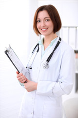 Young brunette female doctor standing with clipboard and smiling at hospital office. Health care, insurance and help concept. Physician ready to examine patient