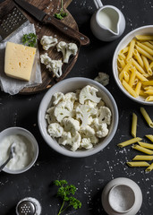 Raw ingredients for cooking pasta with roasted cauliflower - pasta, cauliflower, cheese, cream on dark stone background.