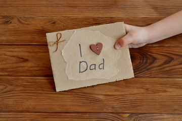 Little child holds a card I love dad. Postcard made of cardboard and wrapping paper, decorated with wooden heart, waxed cord. DIY Father's day or happy birthday card. Gift idea for daddy from daughter