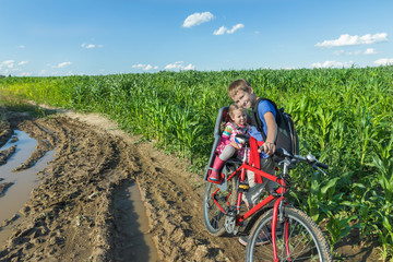 Teenage and preschooler siblings cycling together on summer dirt road in green farm corn field