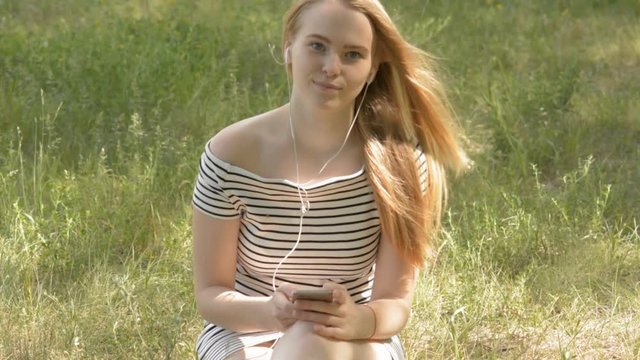 Young woman with headphones and mobile phone outdoors in a park. Her hands pressed buttons on the phone. Girl in a striped dress sitting on the grass
