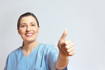 Mature female nurse in a blue scrub smiling and holding her thumb up, light background, lots of empty copy or text space