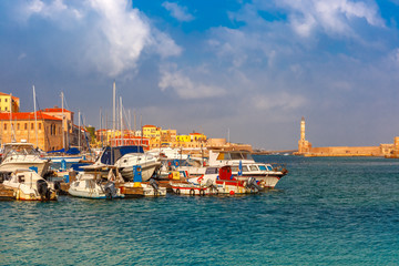 Fototapeta premium Chania Arsenals, the Venetian shipyards, and fishing boats in old harbour of Chania with Lighthouse in sunny and cloudy summer morning, Crete, Greece