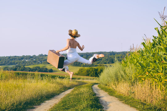 Joyful excited female jumping holding retro suitcase, sunny outdoors country road background. Backview