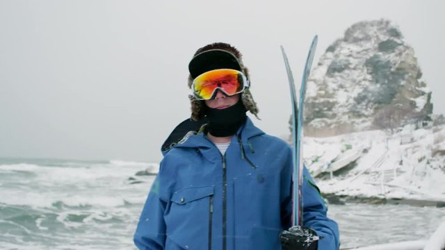 Skier Profile Portrait Holding Skis Wearing Goggles Hat, Jacket, Winter Gear. Looking at Camera Snowy Landscape Mountain Rock, Ocean Waves, Snow Background.