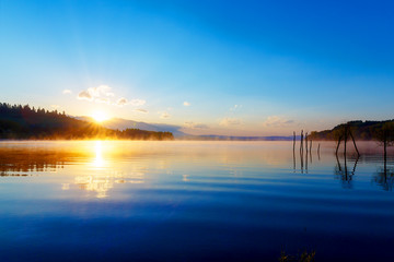 beautiful lake view in morning fog with mystic mountains and trees as leftovers of a mole in gold, purple - blue tones.