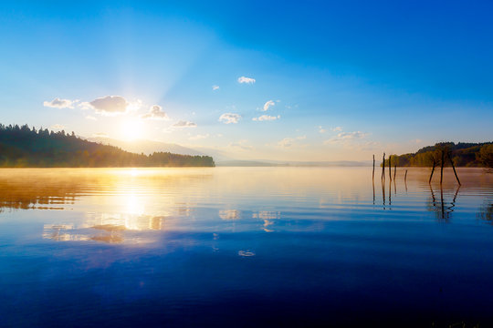 beautiful lake view in morning fog with mystic mountains and trees as leftovers of a mole in gold, purple - blue tones.