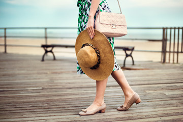 Gorgeous sexy woman wearing summer dress, shoes, pink bag, holding hat and walking on seashore boardwalk at sunset time.