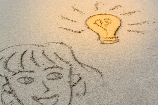A basic female face drawn in the sand with a light bulb shining. 