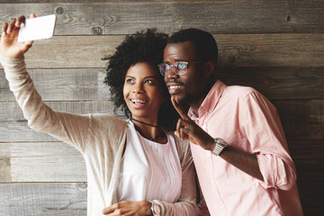 Portrait of lovely African couple having fun and grimacing while taking selfie using mobile phone, posing against wooden wall background. Two happy young people spending time together at a cafe