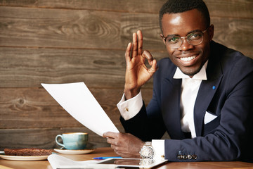Everything is perfect! Portrait of happy cheerful African American executive officer in glasses looking at the camera with confident pleased expression, gesturing 'OK' sign, holding business papers