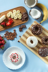 Delicious donuts. Served on the wooden board and white plate. Decorated with strawberry, banana, coconut and almond. Blue background. Silver knife and fork. Vertical image. Top view