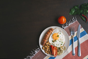 Breakfast set. Pan of fried eggs with sausage, fresh tomato, beans, onion rings, parsley on plate over wooden background, top view, copy space