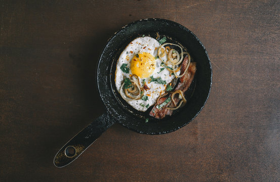 fried egg, bacon, onion rings, parsley - tasty Breakfast or snack, in the old pan on a dark wooden background. Top view.