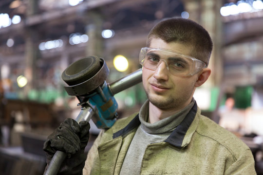 Portrait factory worker in protective glasses with grinding machine on manufacture workshop background