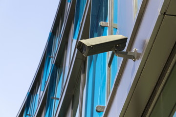 Grey security camera attached to wall blue windows in background