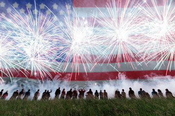 Group of people enjoying fireworks show in a carnival or holiday. People in silhouette and blurred American flag in background.