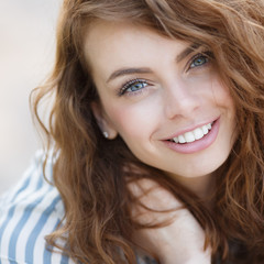Beautiful girl with grey eyes and long curly red hair,a beautiful smile and straight white teeth,wearing earrings,wearing a light striped shirt,posing for a photographer in the summer outdoors