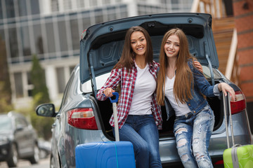two beautiful young brunette woman with long straight hair,nice smile,dressed in blue jeans,a white t-shirt,posing sitting in the open trunk of the gray car,holding a blue and green suitcase on wheels - Powered by Adobe