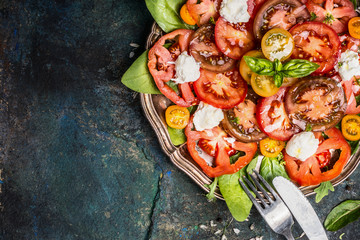 Classic salad with tomatoes and mozzarella in aged metal plate with cutlery on dark rustic background, top view, place for text. Italian food concept