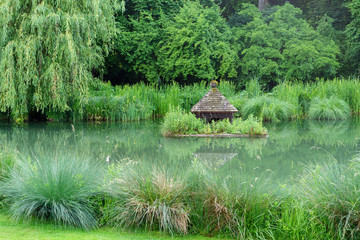 romantischer Blick auf einen von sattem Grün eingefassten, großen Gartenteich mit Entenhütte als...