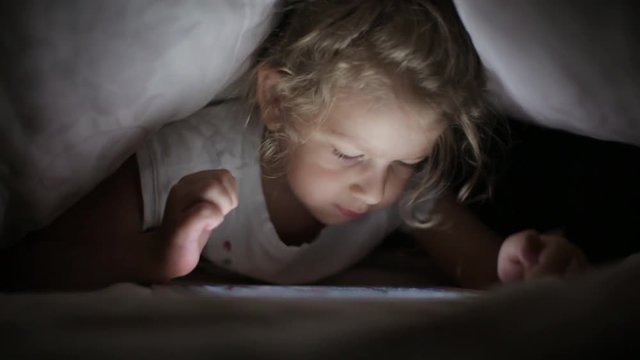 little girl drawing on tablet computer, hiding under the blanket