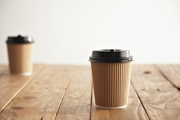 Focused carton paper cup with cap in isolated in center and other unfocused on backside of rustic wooden table
