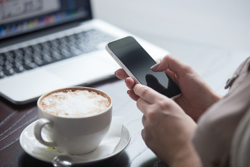 Female hands holding smartphone in coffee shop with laptop on the background. Woman sitting with cup of coffee and using electronic devices in cafe. Close-up