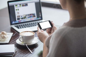 Female hands holding smartphone with empty blank screen for copyspace in coffee shop. Back view. Woman sitting with cup of coffee and syncing files on electronic devices. Close-up