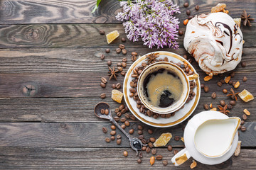 Rustic wooden background with cup of coffee, milk, chocolate meringue and lilac flowers. White vintage dinnerware and spoon. Breakfast at summer morning. Top view, place for text.