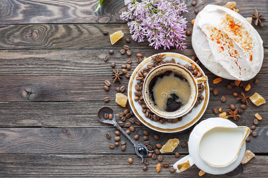 Rustic wooden background with cup of coffee, milk, meringue with peanuts and lilac flowers. White vintage dinnerware and spoon. Breakfast at summer morning. Top view, place for text.