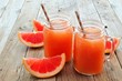 Two mason jar glasses of grapefruit juice with slices and straws on rustic wooden background