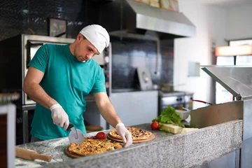 Tuinposter Chef cuts freshly prepared pizza on a wooden substrate. © Аrtranq