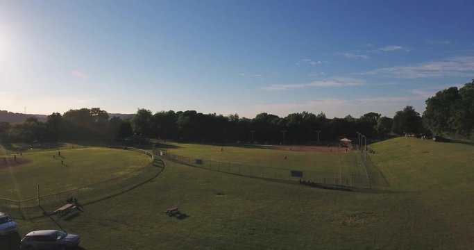 A rising aerial view of kids playing baseball on two public fields at dusk.  	