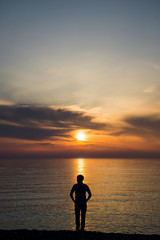 Young man standing at the beach in front of amazing sea view at sunset or sunrise and thinking about his future. Rear view