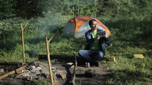 A man taking pictures a fire on the tablet. Against the background is a tent in the forest early in the morning