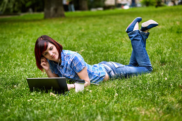 Beautiful young freelancer woman using laptop lying on the grass in the park.Happy smiling girl working online.Studying and learning using notebook computer.Freelance work,business people concept.
