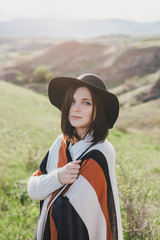 Young beautiful woman traveler wearing hat and poncho relaxing on the top of the hill with mountains and hills around
