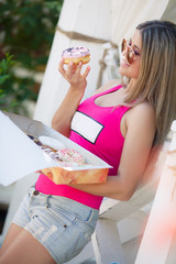 Beautiful blonde girl with long straight hair in sunglasses with brown glasses,dressed in a bright pink shirt and Jean shorts, holding a box of sweet donuts in the shape of a donut