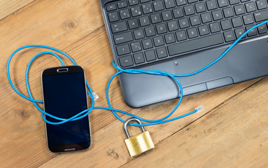 Secure internet system for mobile phone and PC / Smart phone and computer keyboard connected with safety lock and blue wire.
Overhead view with electronic devices on wooden office desk.