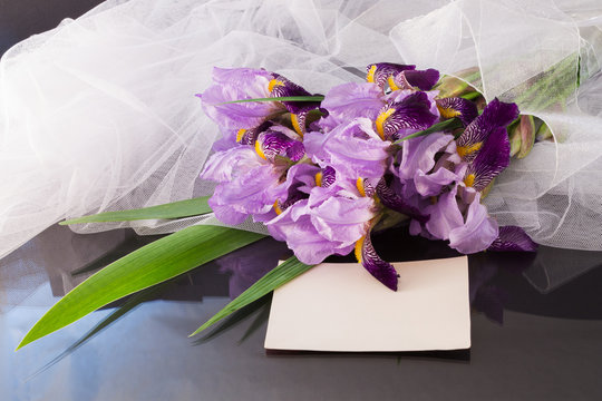 bouquet of purple irises with a veil and a blank sheet of paper on a dark glossy background