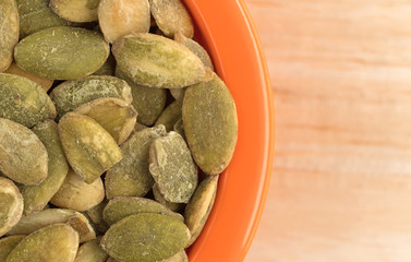 Organic dry roasted pumpkin seeds in a small orange bowl atop a wood table top close view.