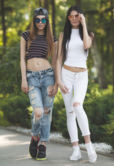 Two attractive young girl friends standing together and posing on camera.Brunette having fun photography at sunset. Summer portrait. Blurred background.