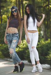 Two attractive young girl friends standing together and posing on camera.Brunette having fun photography at sunset. Summer portrait. Blurred background.