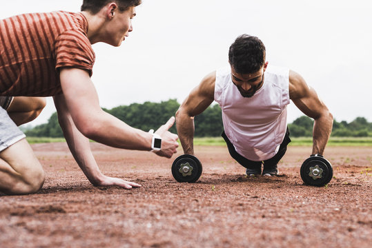 Athlete doing pushups with dumbbells on sports field supported by his training partner