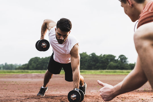 Athlete exercising with dumbbells on sports field supported by his training partner