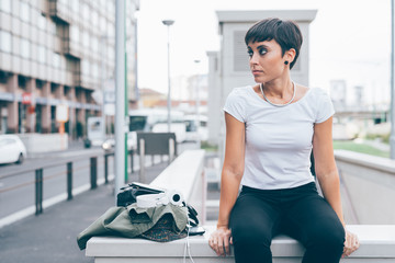 Young beautiful caucasian woman sitting on a small wall outdoor in the city, overlooking pensive,...