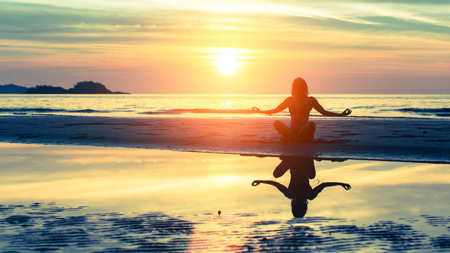Young healthy woman silhouette practicing yoga on the beach at sunset.