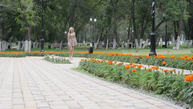 Красивая молодая блондинка идет по аллее летнего парка, кружится и позирует фотографу