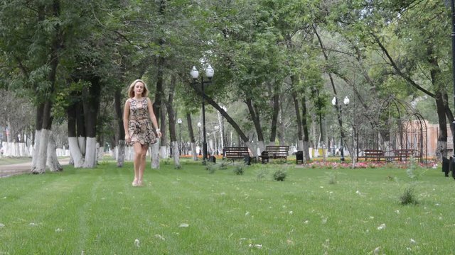 Молодая красивая блондинка идет по траве в летнем парке, кружится и позирует фотографу